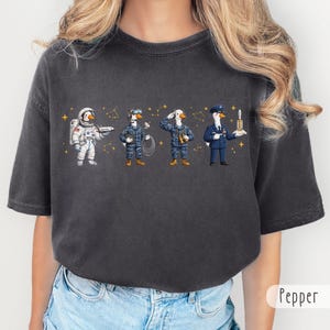 Może przedstawiać: Ciemnoszary t-shirt z nadrukiem graficznym przedstawiającym cztery gęsi w różnych rolach zawodowych: astronautę z promem kosmicznym, technika ze słuchawkami, salutującego żołnierza oraz oficera trzymającego rakietę, wszystko pośród złotych gwiazd.