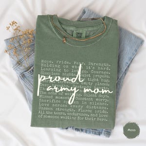 May include: An olive green t-shirt with the words "proud army mom" in a cursive font. The shirt features a list of words and phrases in white, and is folded on top of a pair of blue jeans. A gold necklace rests on the shirt.