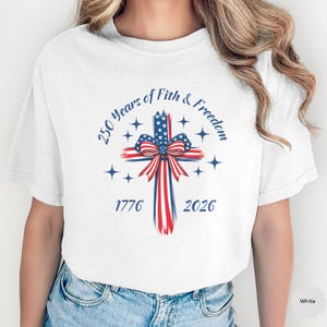 May include: White t-shirt featuring a patriotic cross design. The cross is adorned with the American flag colors and a bow. The text reads "250 Years of Faith & Freedom" with the years 1776 and 2026.