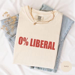 May include: Ivory-colored t-shirt with the text "0% LIBERAL" in red. The shirt is folded on top of a pair of blue jeans. A gold necklace and dried flowers are also visible. The shirt is made by Comfort Colors.