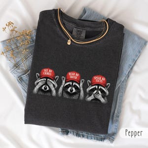 Op de afbeelding: Een antracietgrijs T-shirt met ronde hals en een afbeelding van drie wasberen met rode petten. De eerste wasbeer bedekt zijn ogen met de tekst SEE NO CRIMES, de tweede zijn oren met HEAR NO TRUTH en de derde zijn mond met SPEAK NO FACTS.