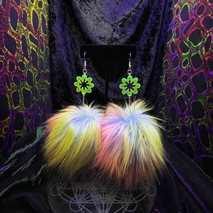 May include: Dangling earrings with neon green flower charms and black centres. Below, fluffy faux fur pom-poms in yellow, pink, blue, and purple hang from the charms. The earrings are displayed against a dark, textured background, with a velvet-like appearance.