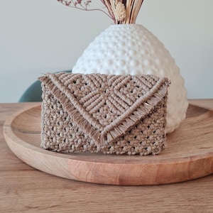 Puede incluir: Clutch de macramé beige hecha a mano, con solapa con flecos. Colocada sobre una bandeja de madera, con un jarrón blanco texturizado y flores secas en el fondo. El bolso tiene un patrón geométrico.