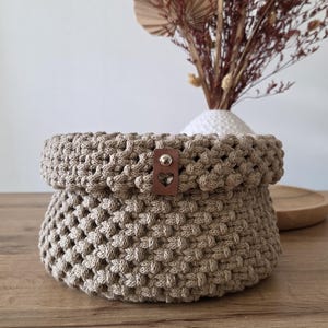 Puede incluir: Una cesta redonda de macramé beige anudada a mano con un patrón tejido texturizado. La cesta presenta una pequeña etiqueta rectangular de cuero marrón sujeta con un remache de metal plateado, que muestra una forma de corazón recortada.