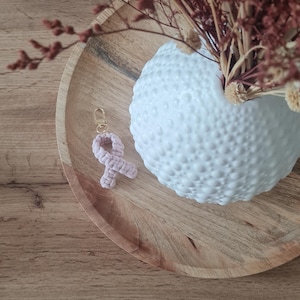 Peut inclure: Un porte-clés ruban en macramé rose clair avec un fermoir mousqueton en métal doré. Le ruban est réalisé avec un motif de cordon noué et est placé à côté d'un vase en céramique blanche texturée rempli de fleurs séchées sur un plateau en bois.
