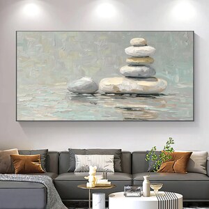 Puede incluir: Una impresión artística en lienzo rectangular representa una pila de piedras lisas y claras en un entorno sereno similar al agua. La obra de arte presenta una paleta de colores neutros con tonos de gris, blanco y beige, creando una estética minimalista y relajante.