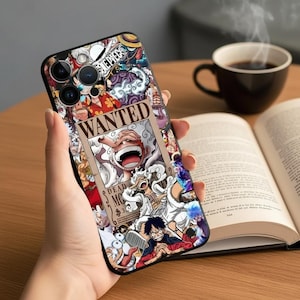 May include: A black phone case featuring a colorful collage of anime characters and the word "WANTED" in a vintage style. The case has a glossy finish and is held in a hand. A cup of coffee and an open book are in the background.