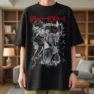 Op de afbeelding: Zwart T-shirt met een grafisch ontwerp uit de anime-serie Death Note. Het ontwerp bevat de tekst "DEATH NOTE" in het rood, samen met illustraties van personages in zwart en wit. Het shirt heeft korte mouwen en een ronde hals.