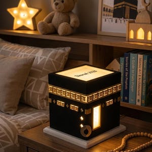 A close-up of the Kaaba night light on a wooden nightstand, featuring gold decorative trim, a glowing door, and the customized text Umrah 2026 on the illuminated top panel.