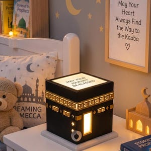 Può includere: Una lampada nera a forma di cubo che ricorda la Kaaba con bordi dorati e il testo "MAY YOUR JOURNEY BE BLESSED" sulla parte superiore. La lampada presenta una luce rettangolare brillante su un lato e un dettaglio circolare argentato. Un rosario in legno è posato accanto.
