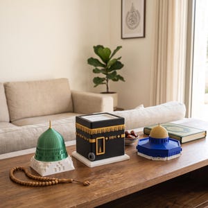 May include: A set of three architectural models including a green domed structure, a black cube with gold calligraphy, and a blue octagonal building with a gold dome, accompanied by a string of brown prayer beads on a wooden surface.
