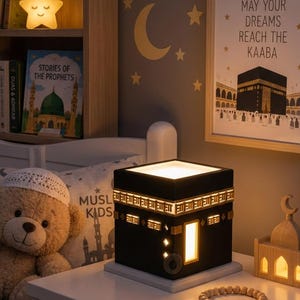 A Kaaba night light rests on a white nightstand next to a teddy bear wearing a white cap, a string of wooden prayer beads, and a framed picture of the Kaaba on the wall.