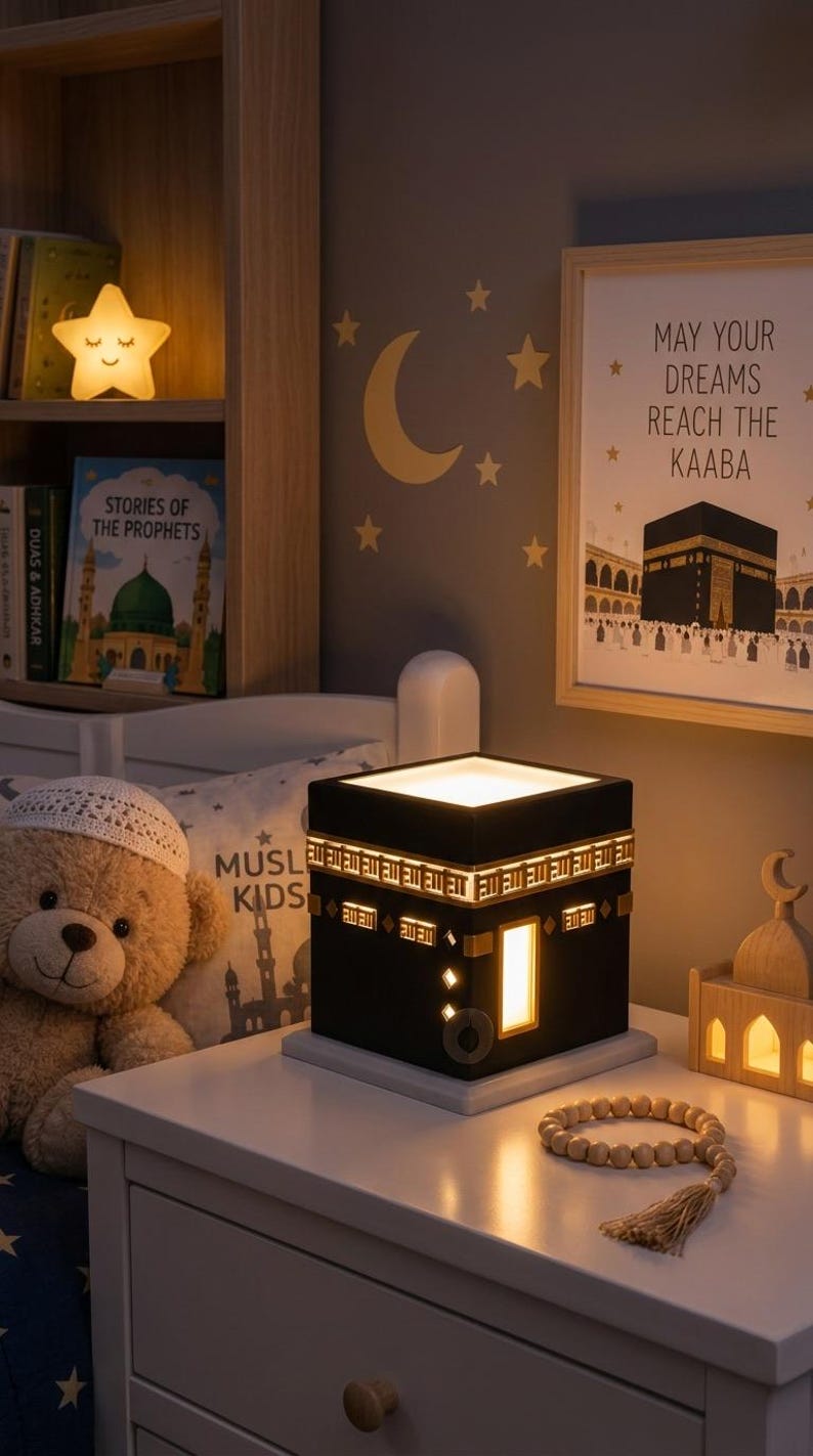A Kaaba night light rests on a white nightstand next to a teddy bear wearing a white cap, a string of wooden prayer beads, and a framed picture of the Kaaba on the wall.