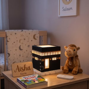 A black Kaaba lamp sits on a wooden nightstand beside a plush camel toy and a personalized wooden plaque.