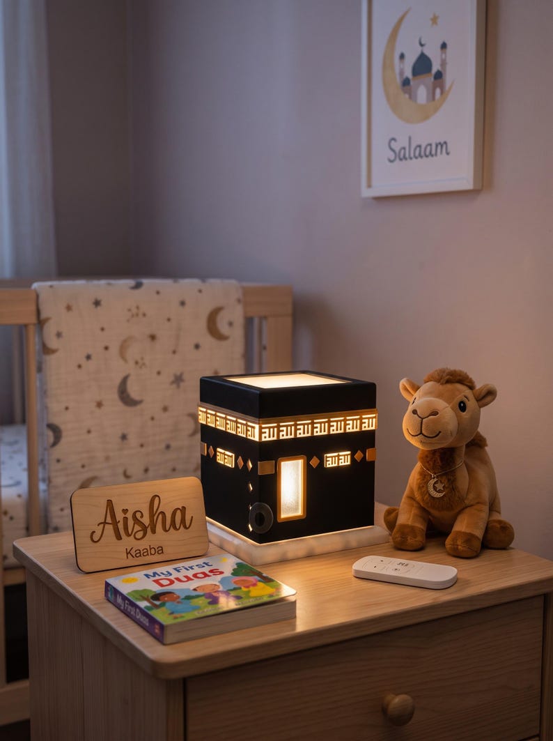 A black Kaaba lamp sits on a wooden nightstand beside a plush camel toy and a personalized wooden plaque.