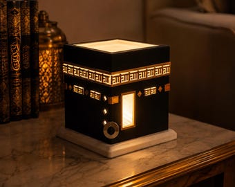 Peut inclure: Une lampe noire carrée en forme de Kaaba, dotée d'une bordure supérieure en calligraphie arabe dorée, d'une ouverture lumineuse rectangulaire verticale et d'accents dorés sur une base blanche. La lampe émet une lueur chaude.