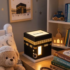 A black cube-shaped Kaaba night light sits on a wooden bedside table in a child room, customized with the text Umrah 2026 on top and glowing warm light.