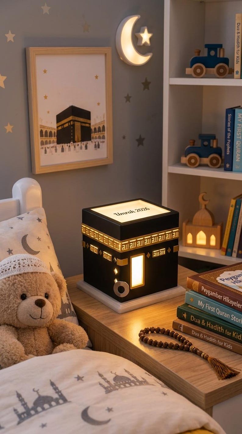 A black cube-shaped Kaaba night light sits on a wooden bedside table in a child room, customized with the text Umrah 2026 on top and glowing warm light.