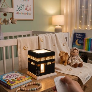 A hand holds a white remote control in front of a glowing Kaaba night light on a wooden dresser.
