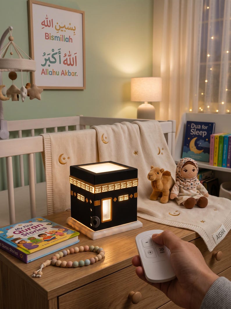 A hand holds a white remote control in front of a glowing Kaaba night light on a wooden dresser.