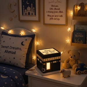 A child bedroom setting features a customized Kaaba night light on a white nightstand, next to books, a teddy bear, a wooden toy sheep, and a pillow reading Sweet Dreams Ahmad.
