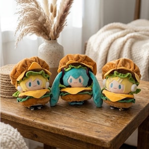 May include: Three plush dolls shaped like burgers, each with a different anime-style character face. The dolls have brown buns, yellow cheese, and green lettuce. The characters have blue, yellow, and green hair. They are sitting on a wooden table.