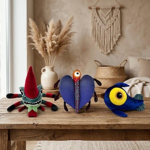 Può includere: Tre peluche a forma di creature stravaganti sono su un tavolo di legno. I giocattoli includono un mostro rosso e verde, una creatura blu a forma di cuore e un pesce blu con un grande occhio giallo. Lo sfondo include un vaso di piante essiccate.