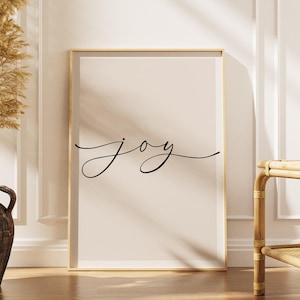 Może przedstawiać: Oprawiony wydruk ze słowem "joy" w eleganckim czarnym skrypcie. Grafika jest umieszczona na neutralnym tle, oprawiona w złoto i oparta o białą ścianę. Po lewej stronie znajduje się dekoracyjny wazon z suszonymi roślinami.