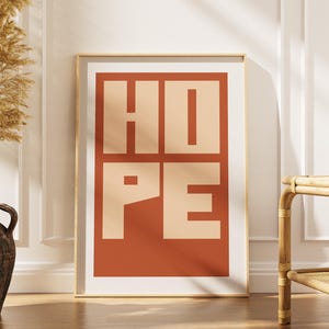 May include: A framed art print with the word "HOPE" in large, block letters. The letters are a light beige color against a terracotta-colored background. The print is in a gold frame and leans against a white wall.