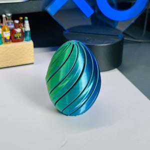 May include: A 3D-printed egg-shaped ornament with a green to blue gradient. The surface has curved, parallel lines, creating a textured effect. The object is displayed on a white surface.