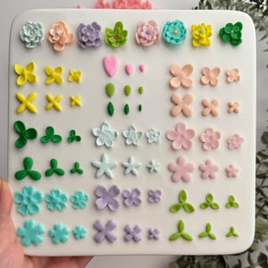 Puede incluir: Una colección de decoraciones florales comestibles y coloridas de varios tamaños y formas. El surtido incluye flores rosas, amarillas, verdes, azules y moradas pastel, junto con hojas verdes y acentos en forma de corazón, todo dispuesto sobre una superficie blanca.