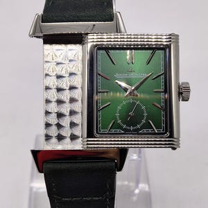 May include: A rectangular wristwatch with a green face and silver case and band. The watch features the Jaeger-LeCoultre logo and a small seconds dial. The watch has a dark green leather strap.