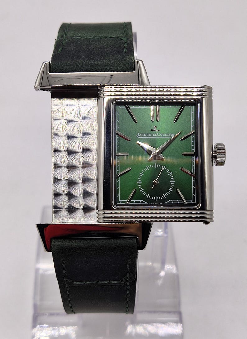 May include: A rectangular wristwatch with a green face and silver case and band. The watch features the Jaeger-LeCoultre logo and a small seconds dial. The watch has a dark green leather strap.