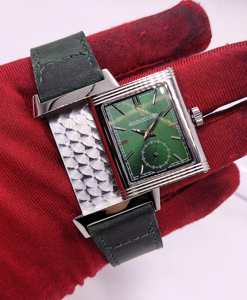 May include: A rectangular wristwatch with a green face and silver-tone case. The watch features a dark green leather strap and a textured silver band. The watch face has the text "Jaeger-LeCoultre" and "Swiss Made".