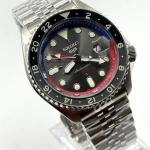 Könnte beinhalten: Eine Seiko GMT Automatik-Uhr aus Edelstahl mit einem schwarzen und grauen Zifferblatt. Die Uhr verfügt über eine drehbare Lünette mit roten, blauen und schwarzen Akzenten und ein Edelstahlarmband. Das Zifferblatt zeigt das Seiko-Logo und den Text "Automatic GMT".