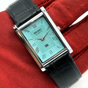 Könnte beinhalten: Rechteckige Seiko Quartz-Uhr mit hellblauem Zifferblatt und silberfarbenem Gehäuse. Die Uhr hat römische Ziffern und ein schwarzes Lederarmband. Das Zifferblatt hat ein strukturiertes Muster und das Seiko-Logo.