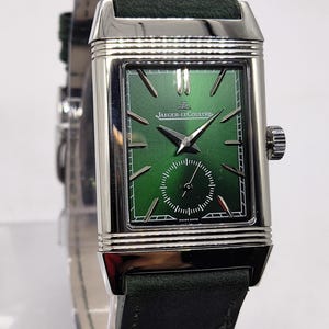 May include: A rectangular wristwatch with a green face and a dark green leather strap. The watch has a silver-coloured casing and the brand name "Jaeger-LeCoultre" is visible on the watch face. The watch has a small seconds dial and a crown on the side.