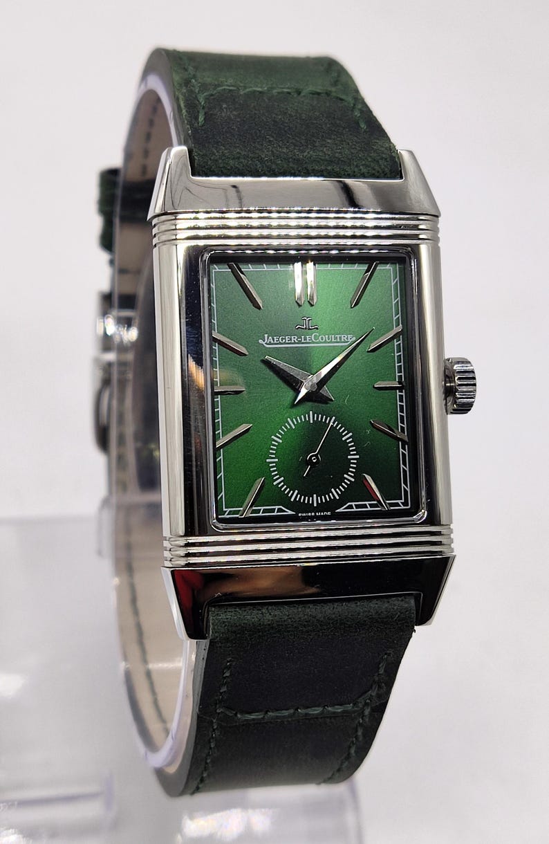 May include: A rectangular wristwatch with a green face and a dark green leather strap. The watch has a silver-coloured casing and the brand name "Jaeger-LeCoultre" is visible on the watch face. The watch has a small seconds dial and a crown on the side.