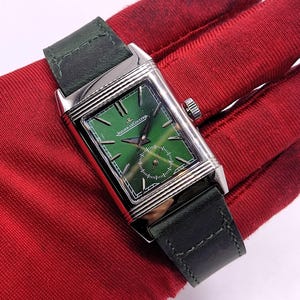 May include: A rectangular wristwatch with a green dial and a silver case. The watch has a dark green leather strap. The watch face features the brand name "Jaeger-LeCoultre".