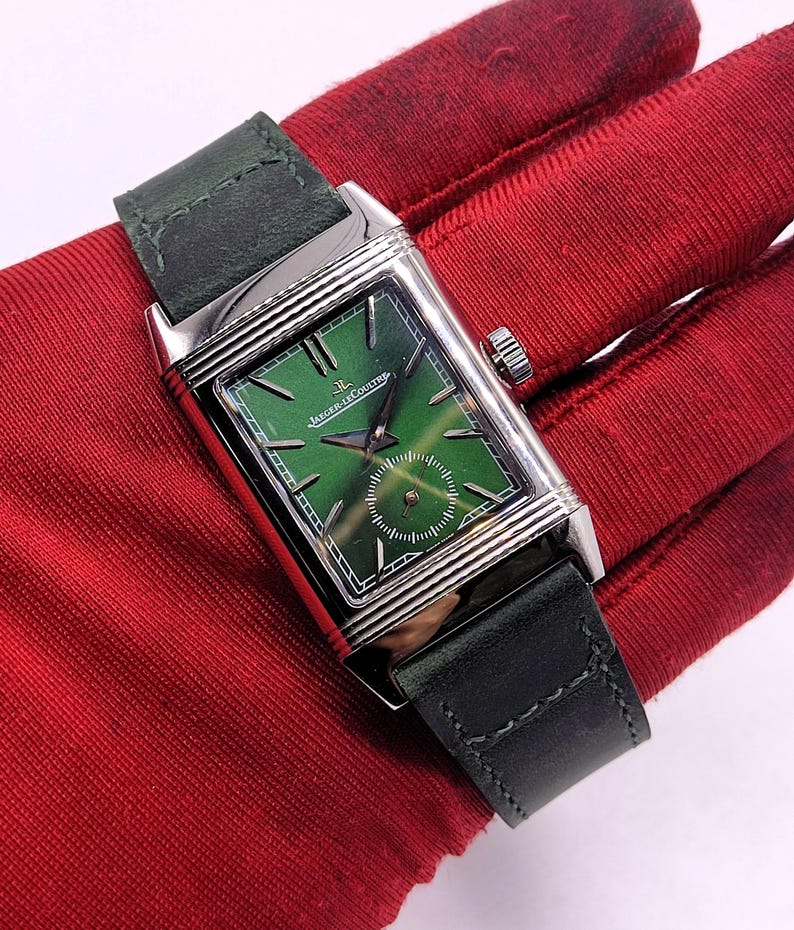 May include: A rectangular wristwatch with a green dial and a silver case. The watch has a dark green leather strap. The watch face features the brand name "Jaeger-LeCoultre".