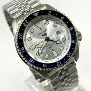 Könnte beinhalten: Eine Seiko 5 GMT Automatik-Uhr aus Edelstahl mit silbernem Zifferblatt, Leuchtmarkierungen und einer blau-schwarzen Lünette. Die Uhr verfügt über eine Datumsanzeige und ein Edelstahlarmband.