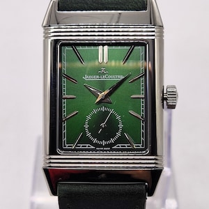 May include: A rectangular, silver Jaeger-LeCoultre watch with a green face and a small seconds dial. The watch has a dark green leather strap. The watch face features the Jaeger-LeCoultre logo and the words "SWISS MADE".