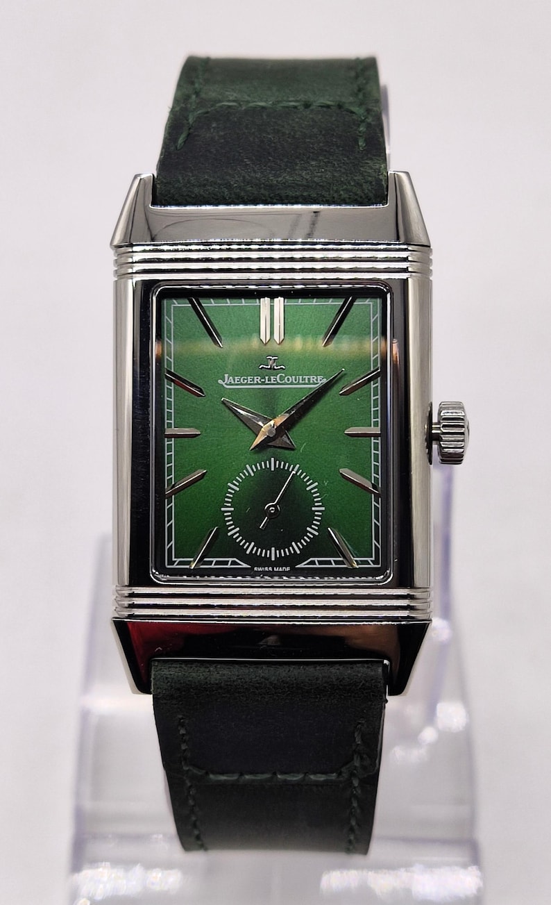 May include: A rectangular, silver Jaeger-LeCoultre watch with a green face and a small seconds dial. The watch has a dark green leather strap. The watch face features the Jaeger-LeCoultre logo and the words "SWISS MADE".