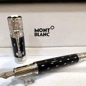 May include: A Montblanc fountain pen with a black barrel and white polka dots. The pen features a silver-colored nib and accents, and the cap is off to the side. The pen is in a white box with the Montblanc logo.