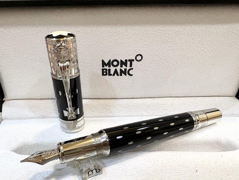 May include: A Montblanc fountain pen with a black barrel and white polka dots. The pen features a silver-colored nib and accents, and the cap is off to the side. The pen is in a white box with the Montblanc logo.