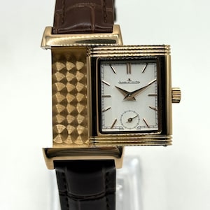 Könnte beinhalten: Eine rechteckige Armbanduhr in Goldoptik mit weißem Zifferblatt und schwarzen Akzenten. Die Uhr hat ein braunes Lederarmband und eine kleine Sekundenscheibe. Auf dem Zifferblatt steht "Jaeger-LeCoultre".