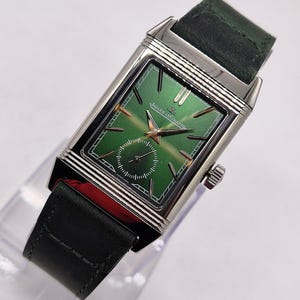 May include: A rectangular wristwatch with a green dial and silver case. The watch has gold hands and markers, a small seconds subdial, and a dark green leather strap. The watch face displays the Jaeger-LeCoultre logo. The watch is a luxury item.