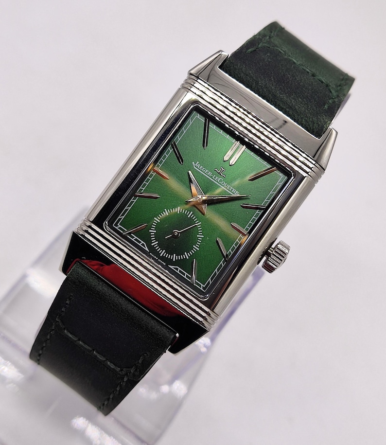 May include: A rectangular wristwatch with a green dial and silver case. The watch has gold hands and markers, a small seconds subdial, and a dark green leather strap. The watch face displays the Jaeger-LeCoultre logo. The watch is a luxury item.