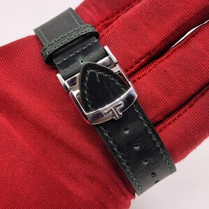 May include: Close-up of a black leather watch strap with a silver buckle. The strap is stitched with green thread and has several holes for adjustment. The buckle features a stylized letter "T".