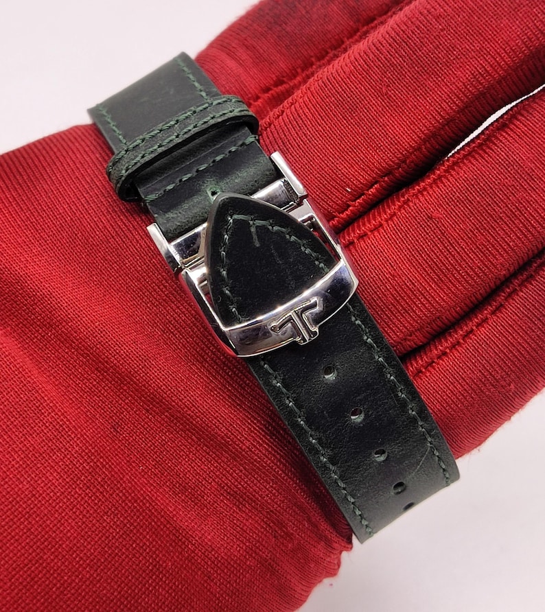 May include: Close-up of a black leather watch strap with a silver buckle. The strap is stitched with green thread and has several holes for adjustment. The buckle features a stylized letter "T".